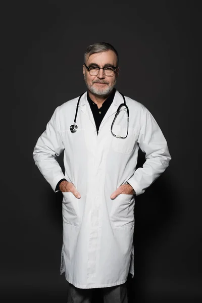 Smiling Senior Doctor Looking Camera While Standing Hands Pockets Dark — стоковое фото