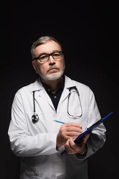 Senior Doctor Eyeglasses Writing Clipboard While Looking Camera Isolated Black — Fotografia de Stock