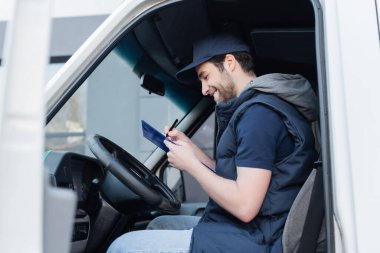 Side view of smiling courier writing on clipboard near steering wheel in car 