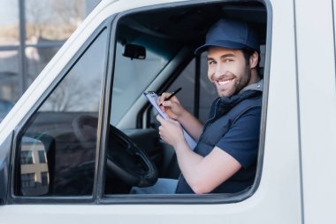 Young courier smiling at camera while writing on clipboard in car 