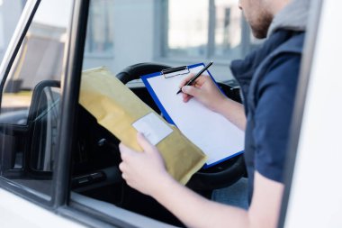 Cropped view of courier writing on clipboard and holding parcel in car 