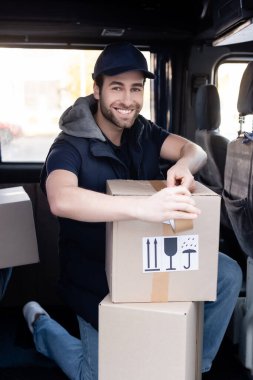 Positive delivery man looking at camera while applying adhesive tape on box in car 