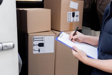 Cropped view of courier writing on clipboard near blurred carton boxes in auto 