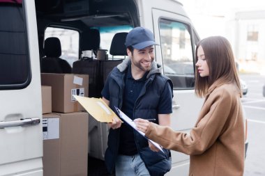 Smiling delivery man holding parcel near woman writing on clipboard and car outdoors 