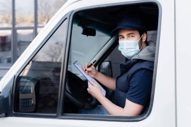 Courier in medical mask holding clipboard and looking at camera in car 