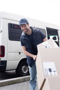 Smiling courier looking at carton boxes with signs near auto outdoors 