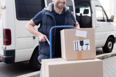 Cropped view of delivery man holding cart with cardboard boxes on urban street 