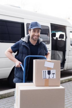 Cheerful delivery man holding cart with packages near blurred car outdoors 