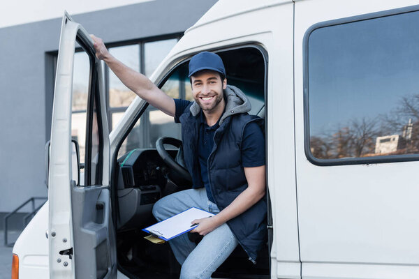 happy delivery man looking at camera while holding clipboard and parcel in auto 