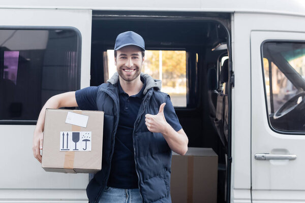 Smiling courier showing like and holding carton box near car outdoors 