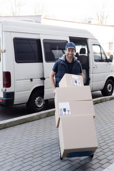 Smiling courier in cap holding cart with boxes near car outdoors 