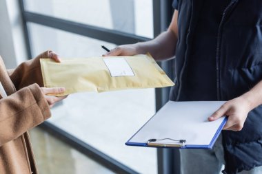 Cropped view of courier holding clipboard and giving parcel to woman in hallway 