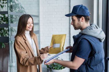 Positive woman holding parcel and writing on clipboard near delivery man in hallway 