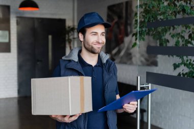 Smiling courier in uniform holding carton box and clipboard in hallway 
