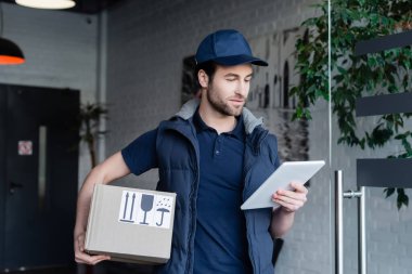 Delivery man in uniform holding carton box and using digital tablet in hallway 