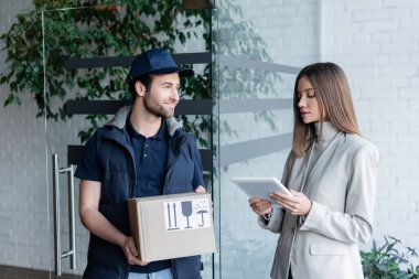 Young woman holding digital tablet near smiling delivery man with carton box in hallway 