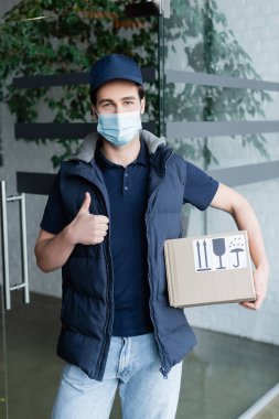 Courier in medical mask holding box with symbols and showing like in hallway 