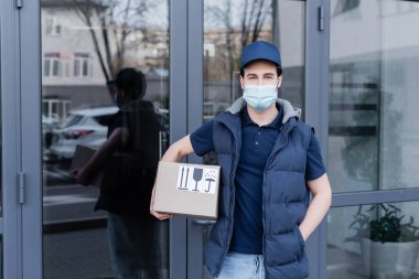 Young delivery man in medical mask holding cardboard box and looking at camera near building outdoors  clipart