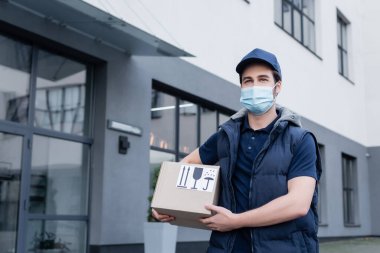 Delivery man in medical mask holding carton box with signs on urban street 