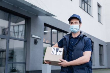 Delivery man in medical mask holding carton box with signs on urban street  clipart