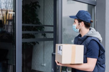 Courier in protective mask holding carton box near door of building outdoors 