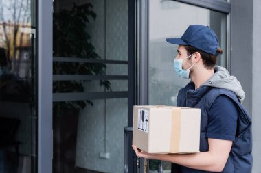 Courier in protective mask holding carton box near door of building outdoors  clipart