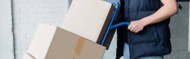 Cropped view of courier holding cart with cardboard boxes in hallway, banner 