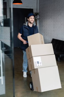 Positive courier standing near cart with packages in hallway  clipart