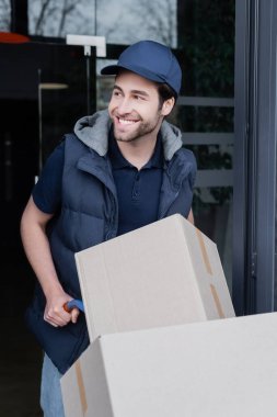 Smiling delivery man looking away near car with carton boxes and building outdoors 