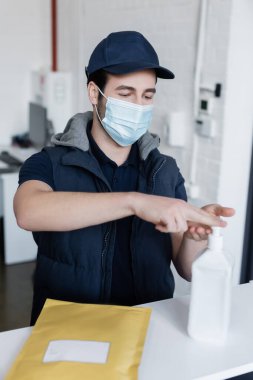 Courier in medical mask applying hand sanitizer near parcel on reception in office 