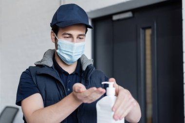 Delivery man in medical mask applying blurred hand sanitizer in office 