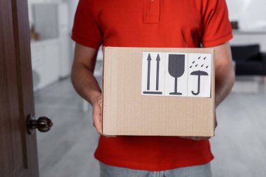 Cropped view of courier holding carton box with symbols in hallway  clipart