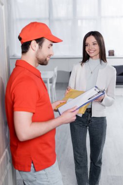 Positive woman taking parcel from delivery man with clipboard in hallway 