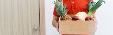Cropped view of courier holding box with fresh fruits and vegetables near door in hallway, banner  clipart