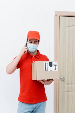 Courier in medical mask talking on smartphone and holding cardboard box in hallway  clipart