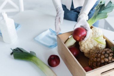 Cropped view of woman in latex gloves holding apple near fresh food and antiseptic at home 