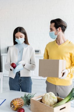 Man in medical mask holding carton box near girlfriend cleaning apple with rag at home 