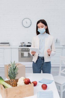 Woman in medical mask holding digital tablet near fresh food and carton box in kitchen 