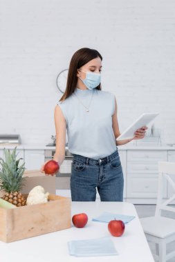 Woman in medical mask holding apple near boxes and using digital tablet in kitchen 