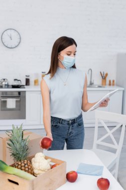 Woman in medical mask using digital tablet and taking apple from wooden box in kitchen 