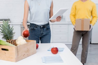 Cropped view of woman using digital tablet near fresh food and boyfriend with cardboard box at home 