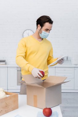 Man in medical mask and latex gloves using digital tablet and holding apple near carton box at home 