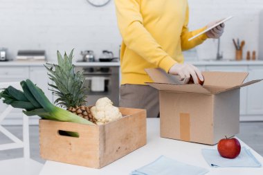 Cropped view of man in latex gloves using digital tablet and taking apple from carton box in kitchen 