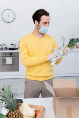 Young man in medical mask cleaning leek near fresh food and digital tablet in kitchen 