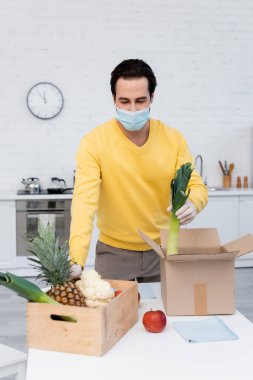 Young man in medical mask holding leek near fresh food and rags at home 