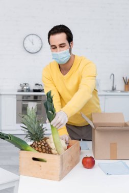 Man in protective mask taking leek from box with food near rags in kitchen 