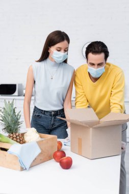 Couple in medical masks looking at carton box near fresh food and rags in kitchen 