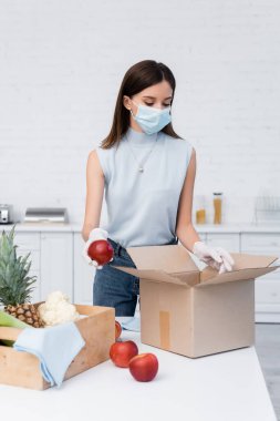 Woman in latex gloves and medical mask taking apple from carton box near rags in kitchen 