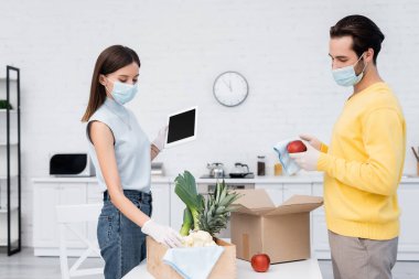 Woman in medical mask holding digital tablet near boyfriend cleaning apple and boxes in kitchen 