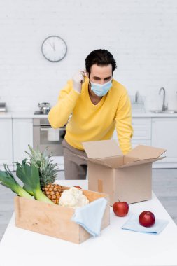 Man wearing medical mask near fresh food and boxes in kitchen 